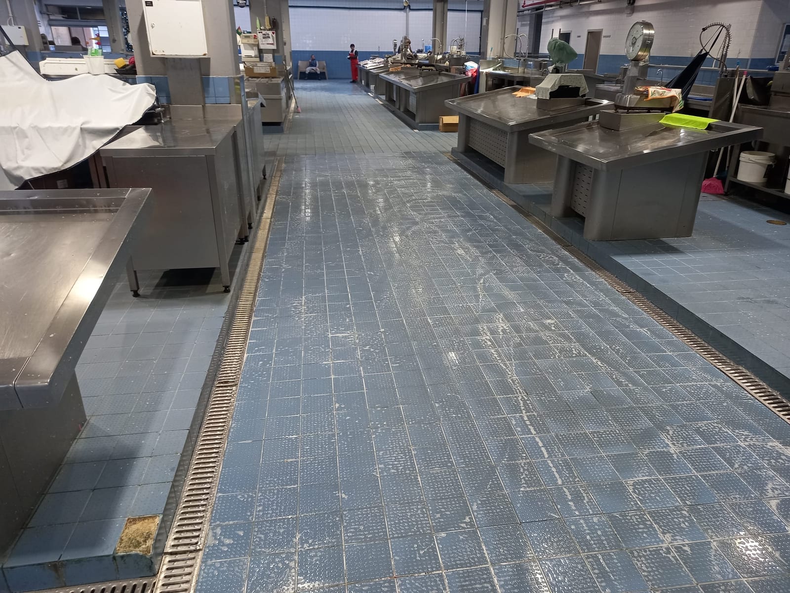 Corredor entre bancas de inox com pavimento azul molhado e restos de espuma durante a lavagem.