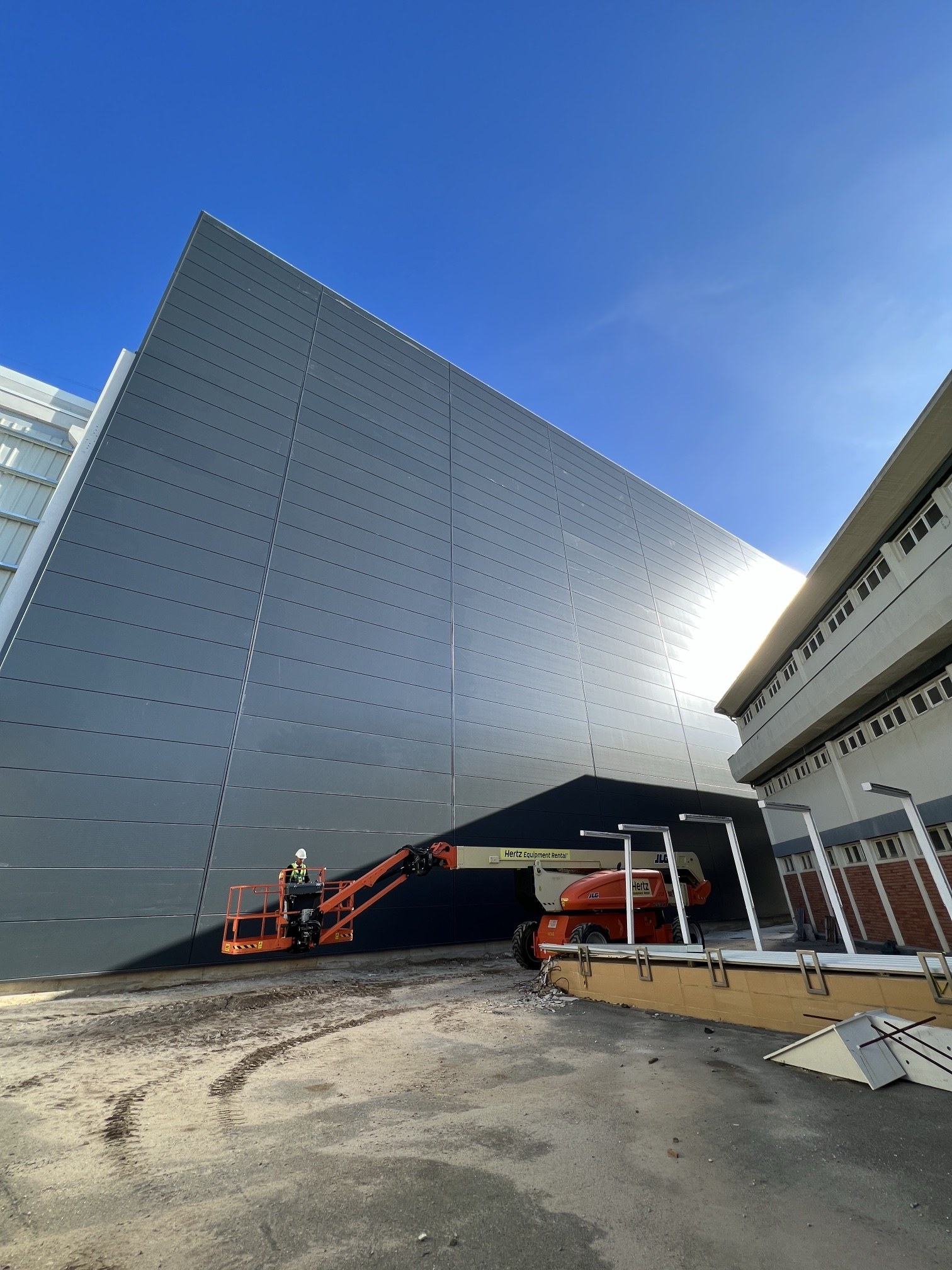 Técnico com capacete e colete numa plataforma elevatória articulada junto a uma fachada industrial em chapa escura em Aveiro